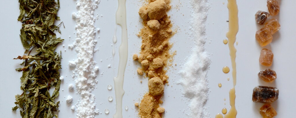 Hier haben wir verschiedene Zuckersorten abgebildet - ihr seht von links nach rechts getrocknete Stevia-Bl&auml;tter, raffinierten Zucker in Form von Puderzucker, Agavendicksaft, Vollrohrzucker, Xylit, Ahornsirup und brauner Zucker als Kandis, also nicht fein gemahlen, sondern grob zersto&szlig;en.